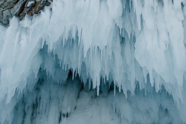 A dramatic close-up of sharp icicles hanging at a cave entrance in a winter setting.