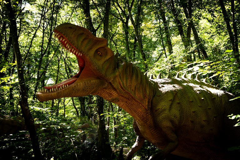 A lifelike dinosaur model roars in a vibrant, sunlit forest setting.