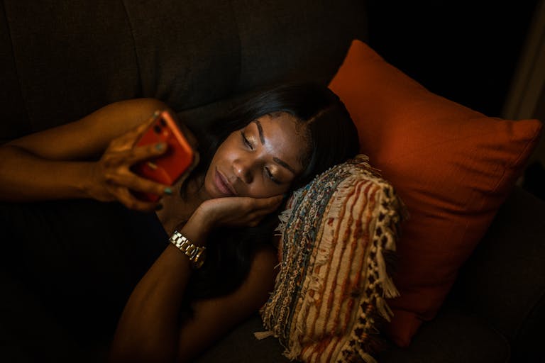 A woman comfortably lying on the couch browsing her smartphone at night with cozy pillows.