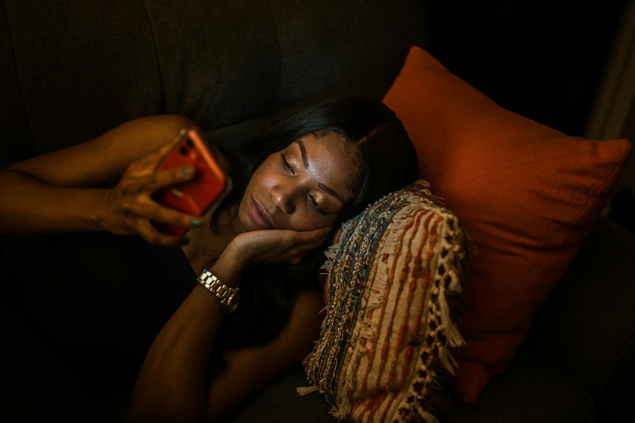 A woman comfortably lying on the couch browsing her smartphone at night with cozy pillows.