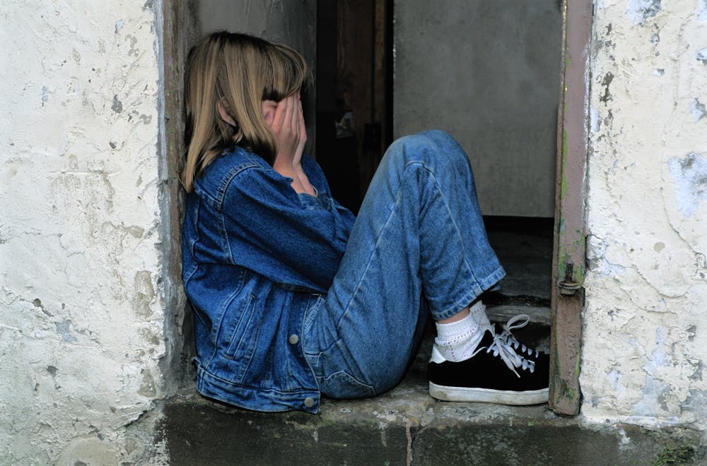 The Famous “Unhappiness Hump” Has Disappeared – Youth Mental Health is Now at Its Worst A young girl in denim sits alone in a doorway looking distressed, conveying loneliness.