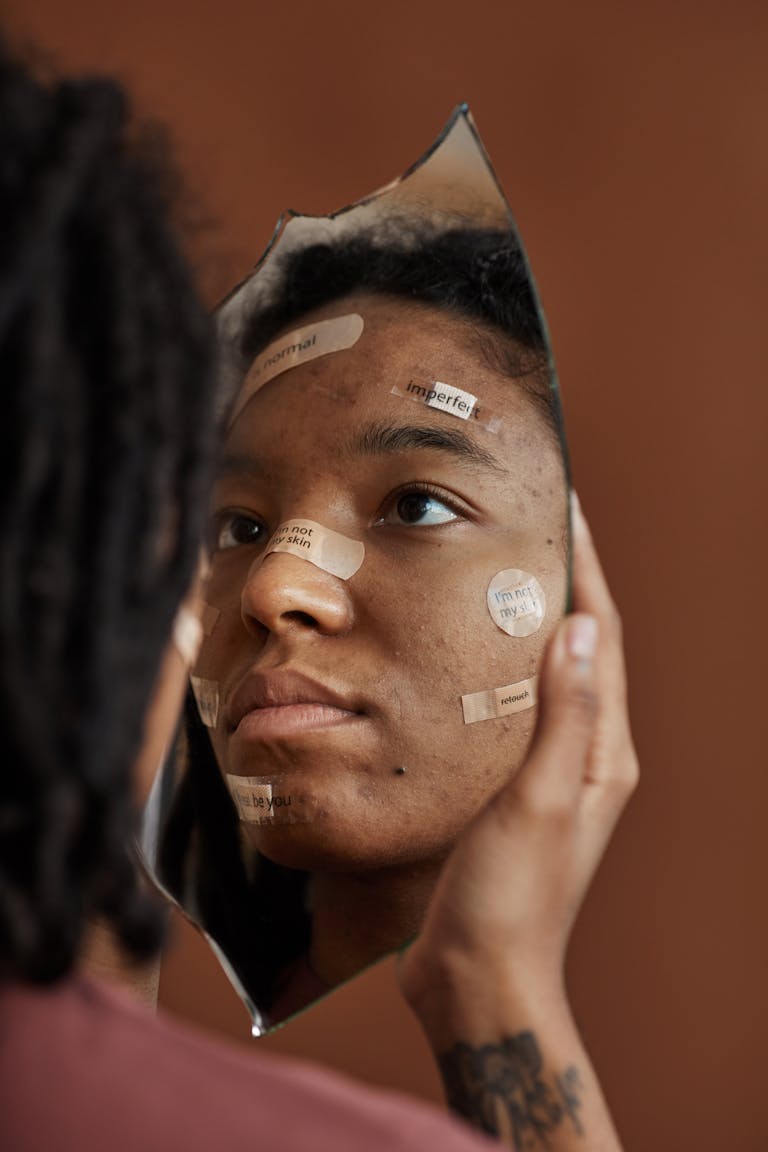 A young woman observes her reflection in a broken mirror with positive affirmations on band-aids.