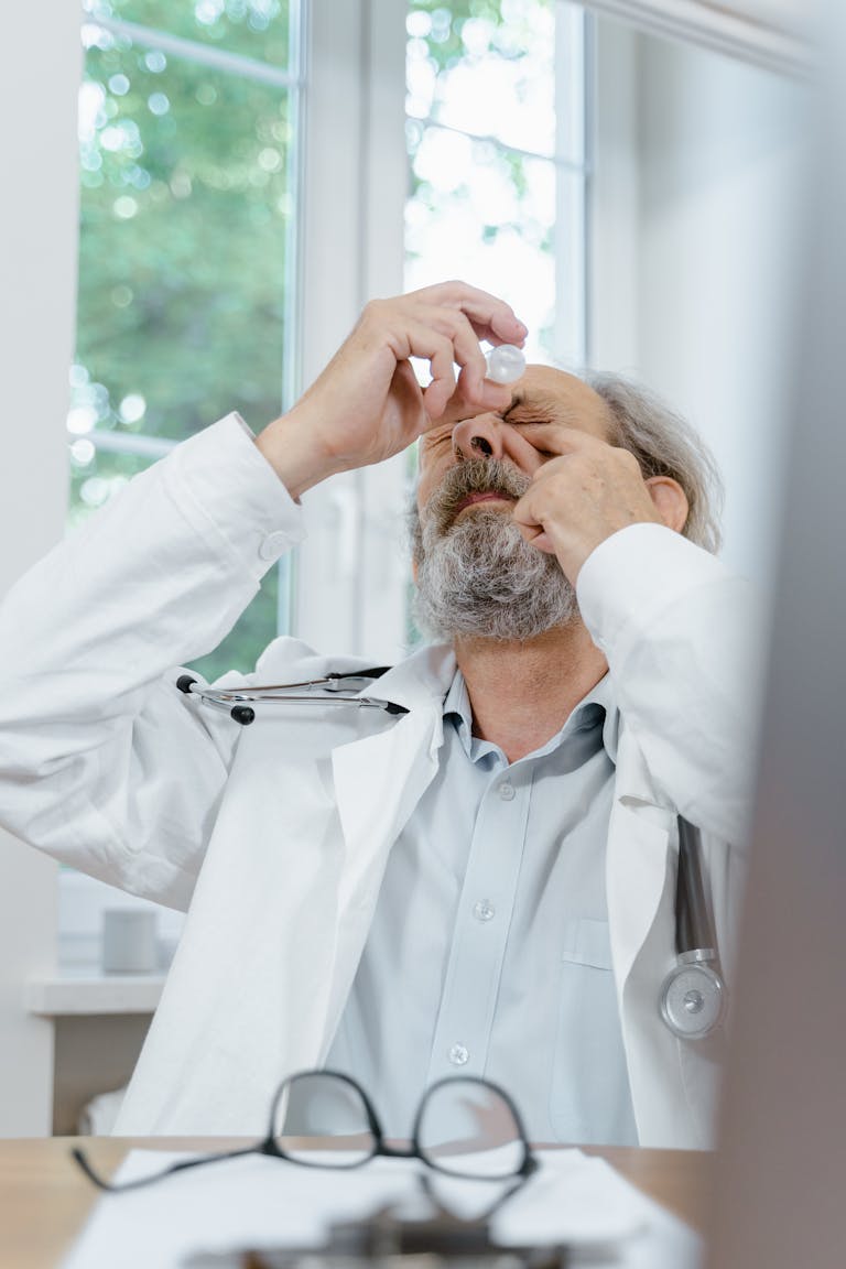 Senior male doctor in white coat applying eyedrops, sitting in a well-lit clinic.