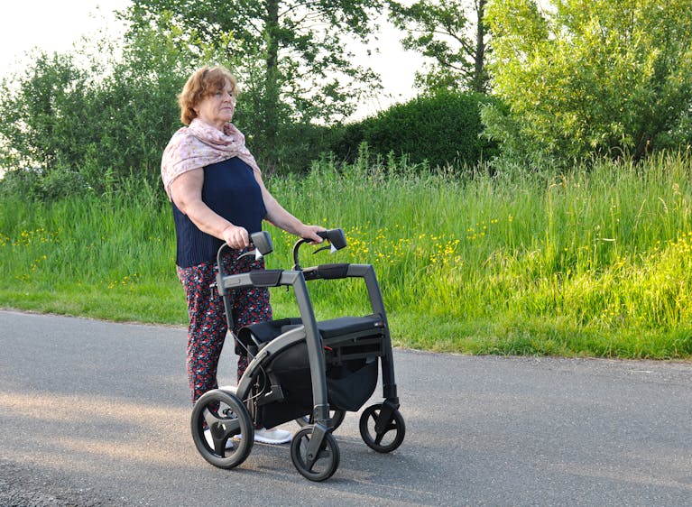 Senior woman enjoying a sunny day outdoors with a rollator walker, surrounded by green nature.