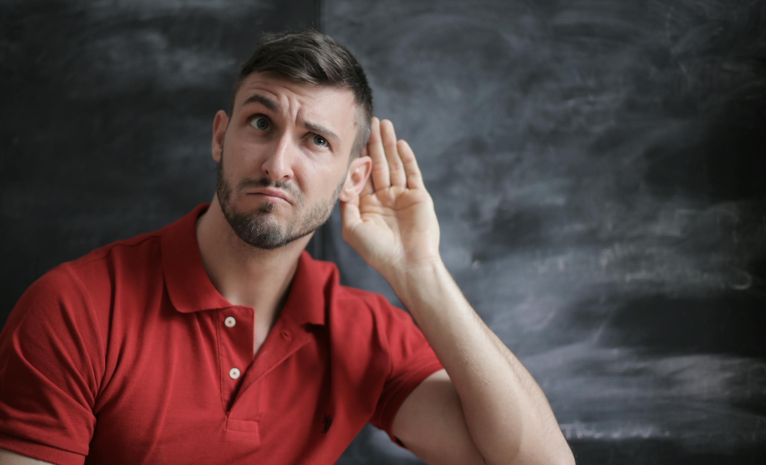 IQ and the Ability to Hear in Noisy Environments: What a New Study Reveals Thoughtful man listening intently against a chalkboard background in a red polo shirt.