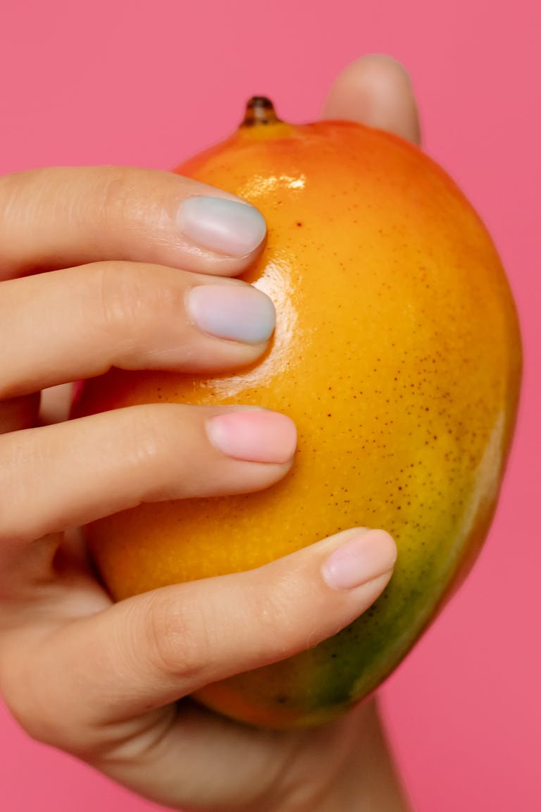 A close-up shot of a hand with painted nails holding a ripe mango against a pink background.