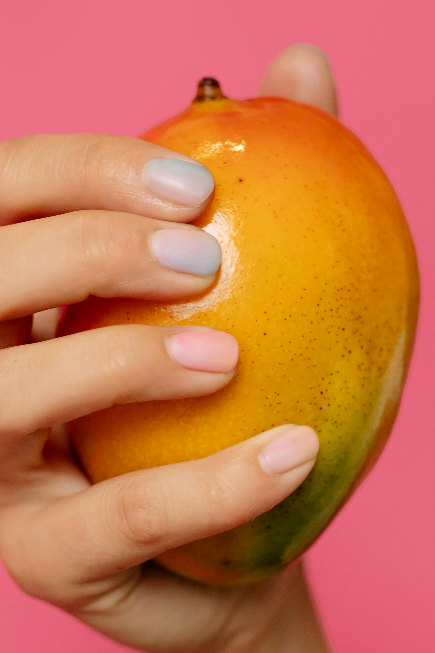 A close-up shot of a hand with painted nails holding a ripe mango against a pink background.