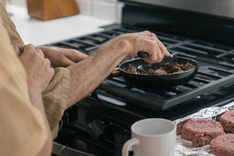 Close-up of hands preparing meat in a frying pan on a stovetop, home cooking scene.
