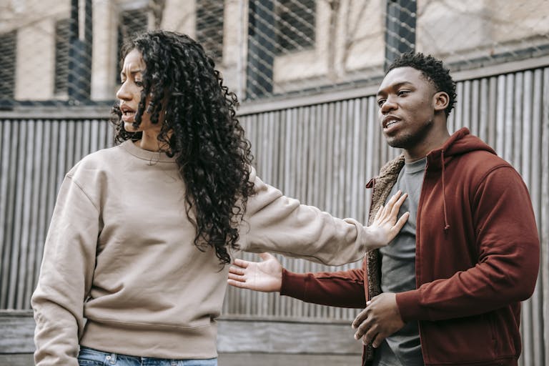 A couple in casual clothing appears to be in a disagreement outdoors with grey fencing background.