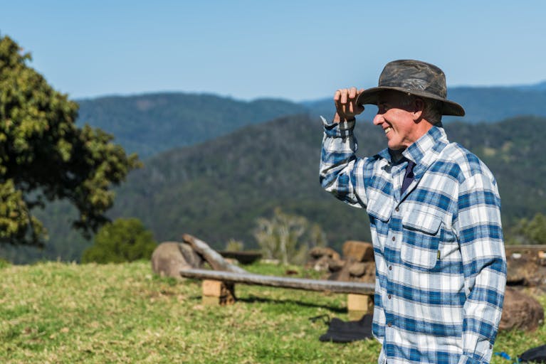A smiling farmer in plaid flannel enjoys a sunny day on a lush green hillside.
