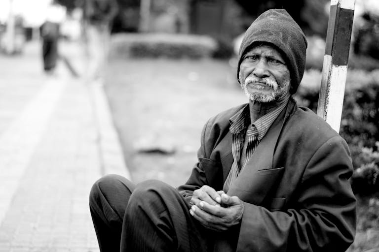 A black and white portrait of an elderly man sitting on the streets, expressing hardship and resilience.