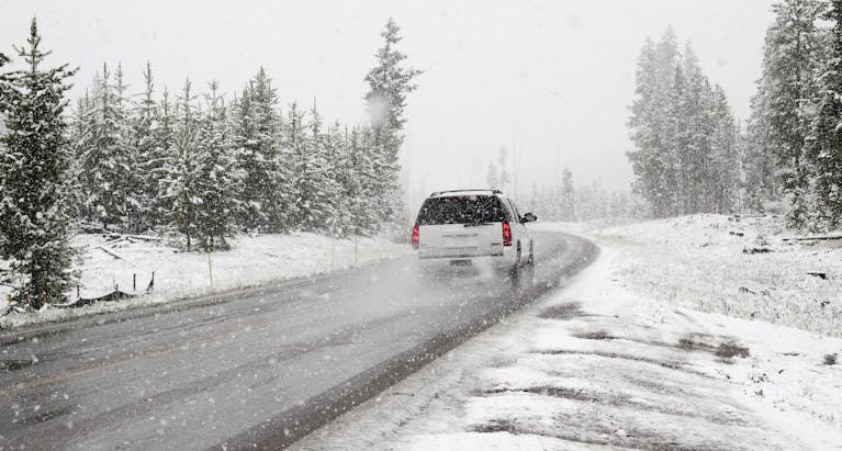 A car drives on a snowy road surrounded by trees during a snowstorm, creating a wintry scene.