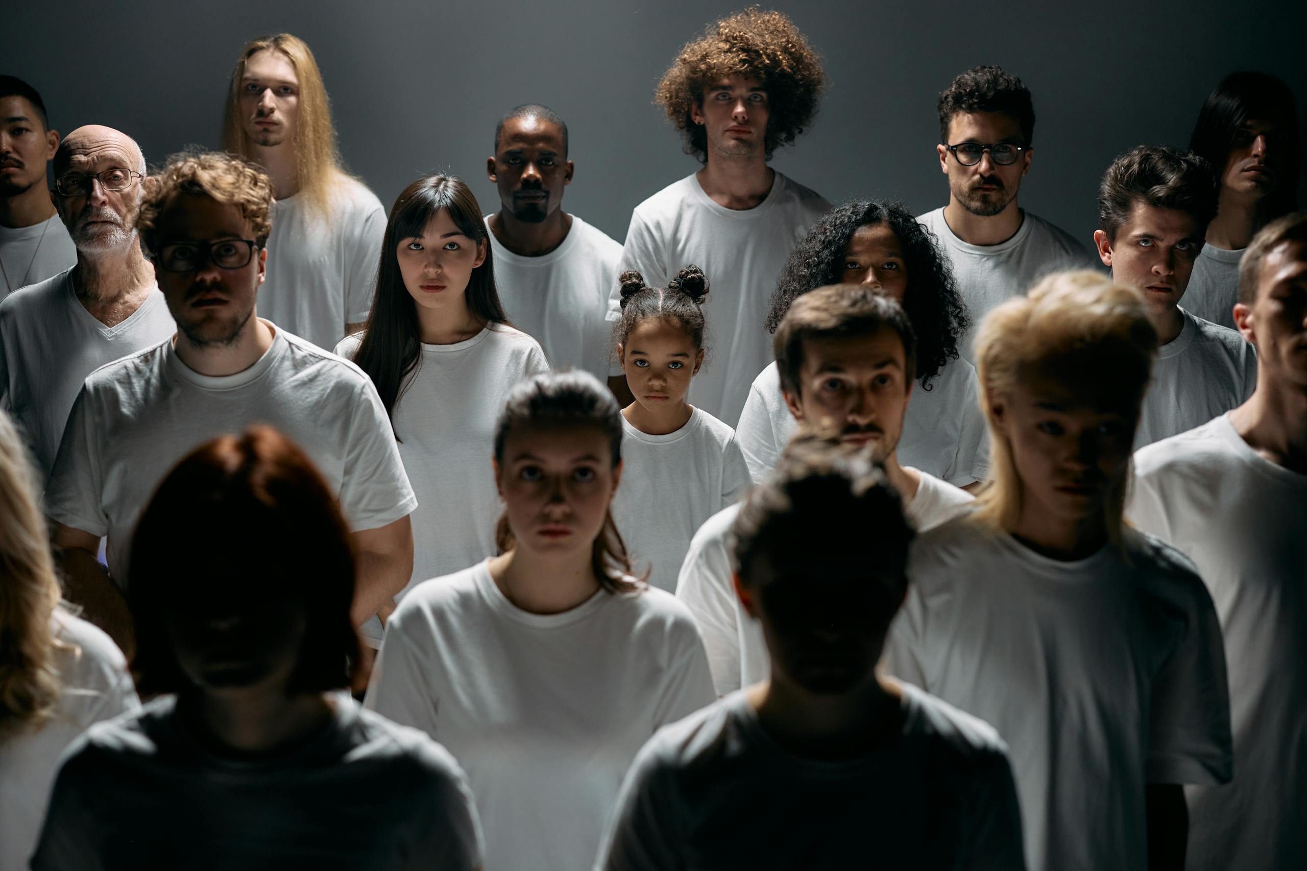 A diverse group of individuals stands together, emotionless, in a studio setting.