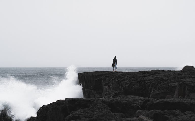 A lone person stands on a rocky cliff in Iceland with waves crashing below under a gloomy sky.