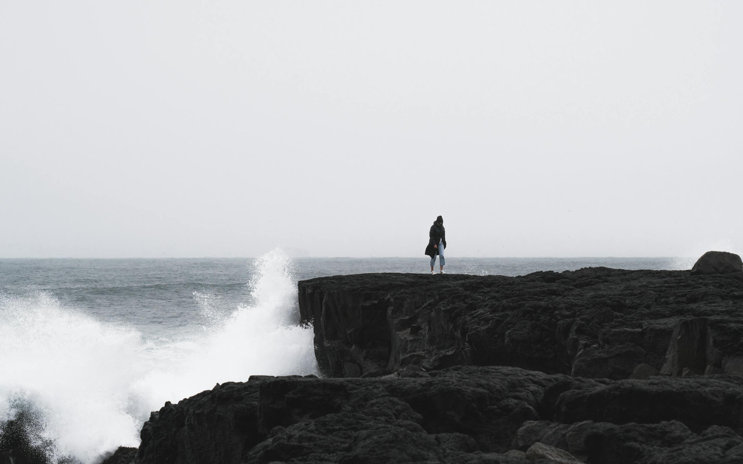 A lone person stands on a rocky cliff in Iceland with waves crashing below under a gloomy sky.