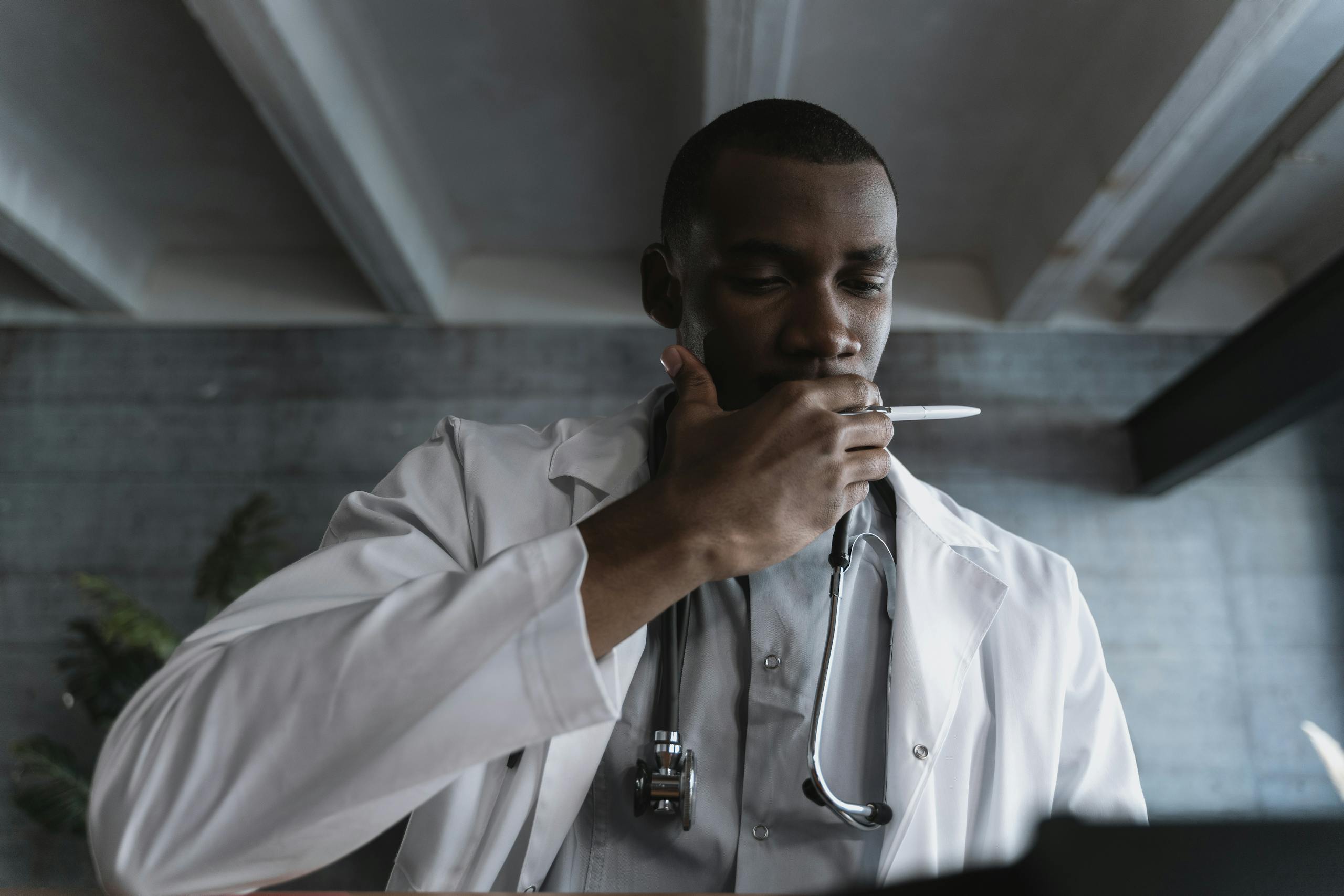 A male doctor in deep thought, holding a pen and wearing a stethoscope indoors.