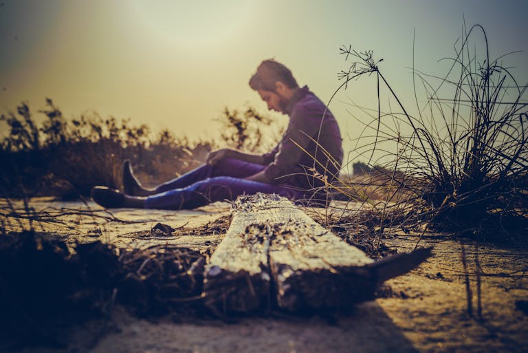 A man sits alone on sandy terrain at sunset, capturing solitude in nature's calm.
