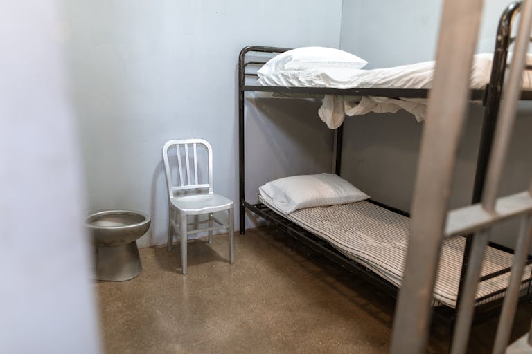 A stark prison cell featuring a bunk bed, metal chair, and toilet.
