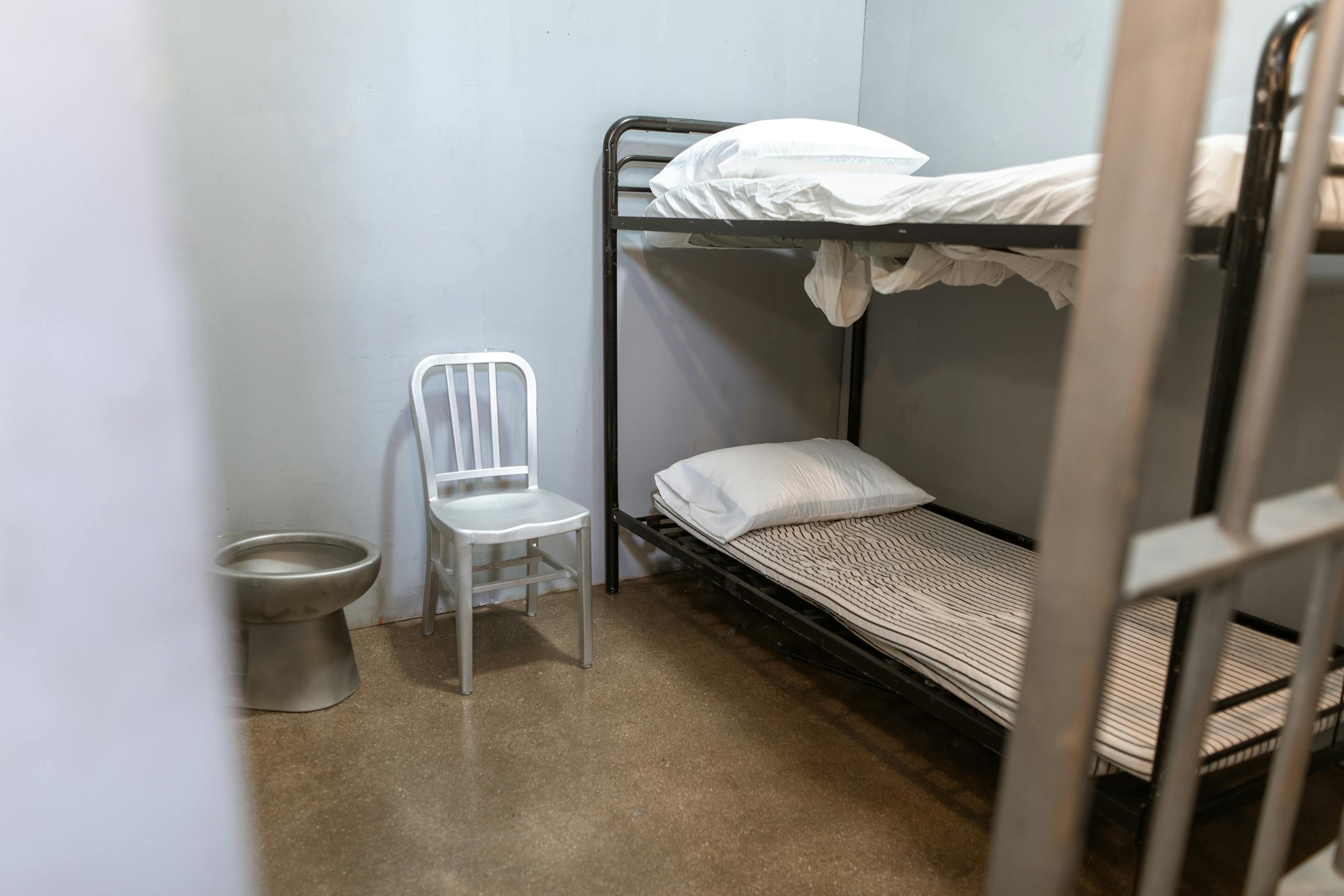 A stark prison cell featuring a bunk bed, metal chair, and toilet.