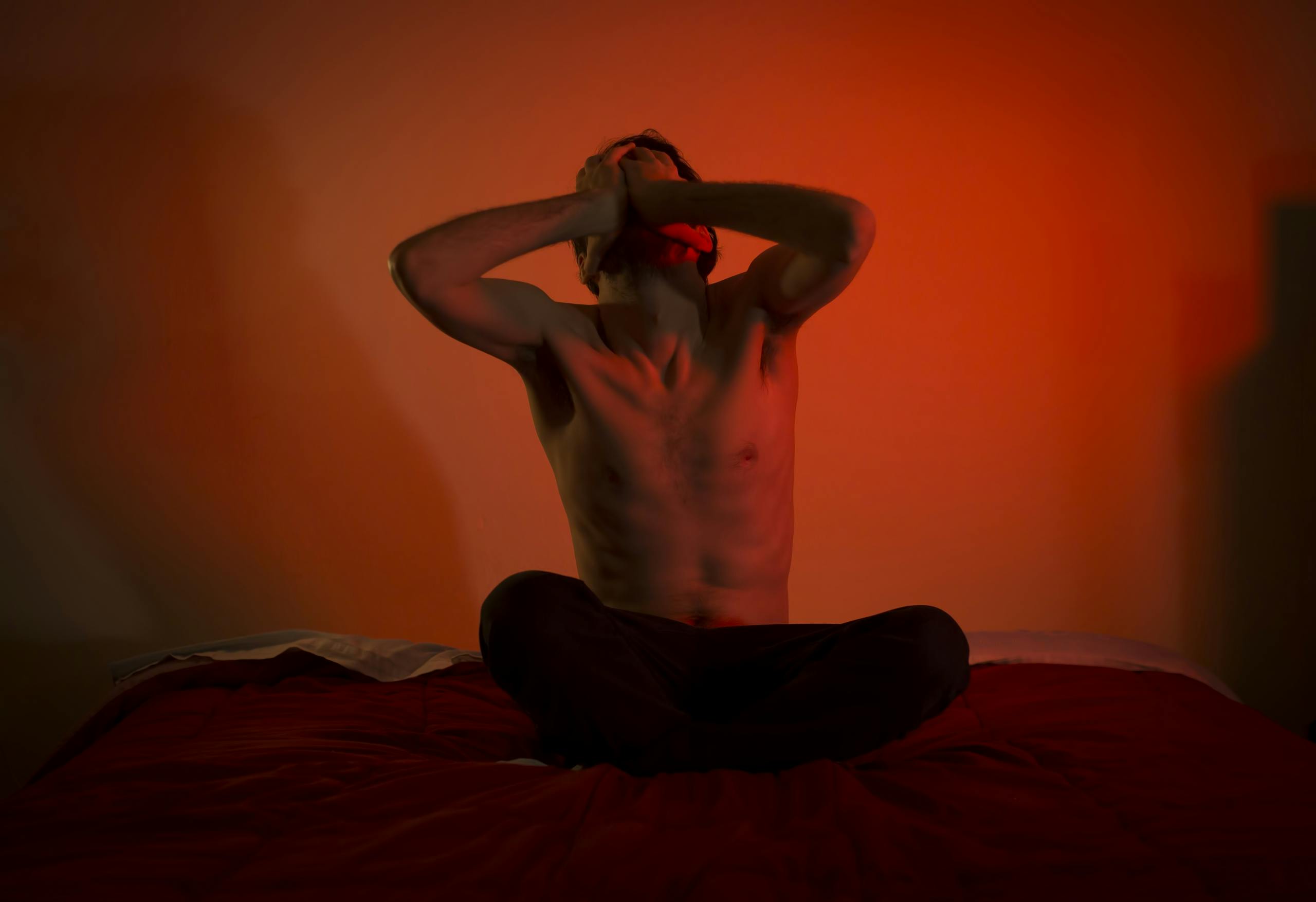 A topless man showing emotional distress sitting on a bed in a dimly lit room.