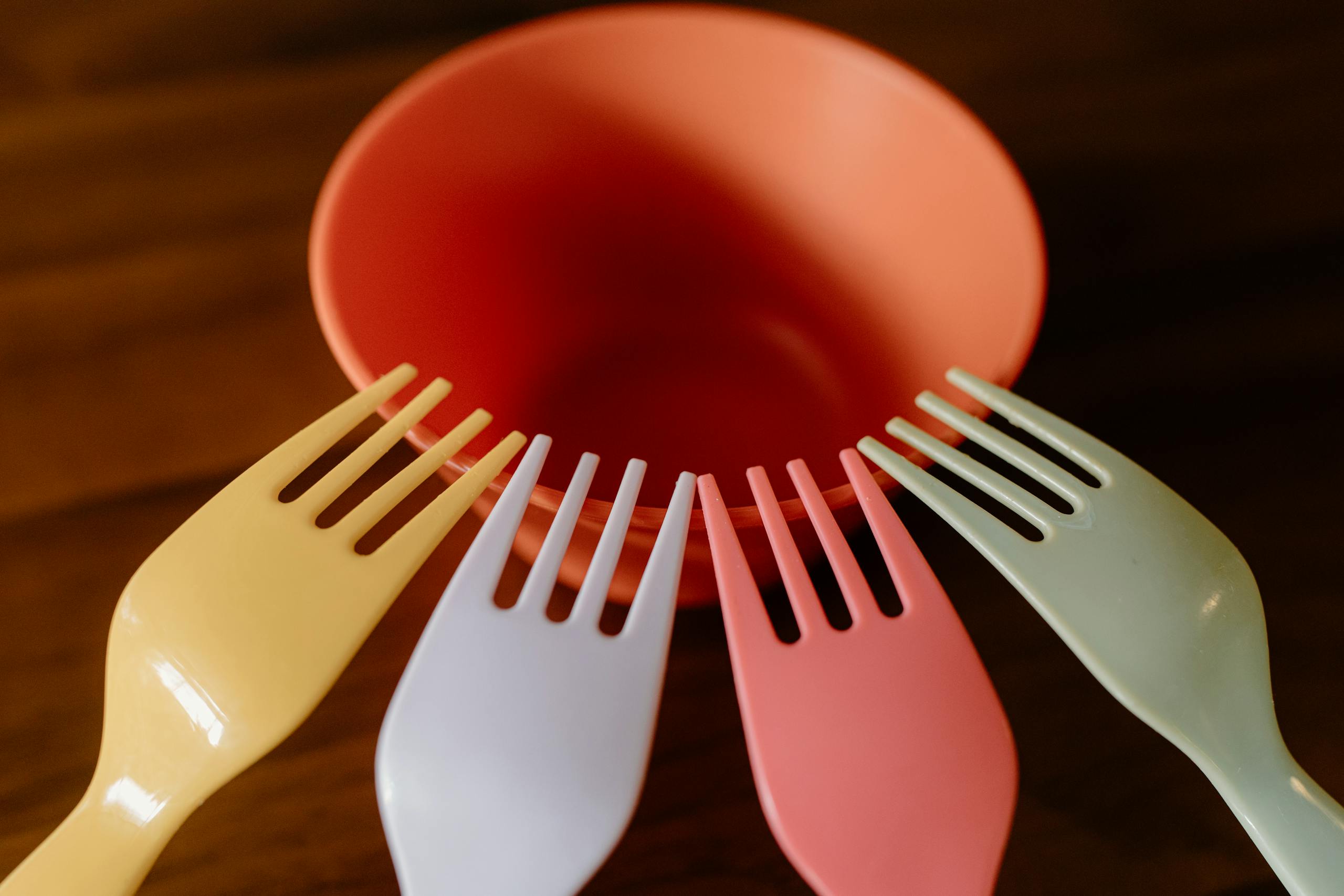 A vibrant close-up of colorful plastic forks surrounding an orange bowl on a wooden table.