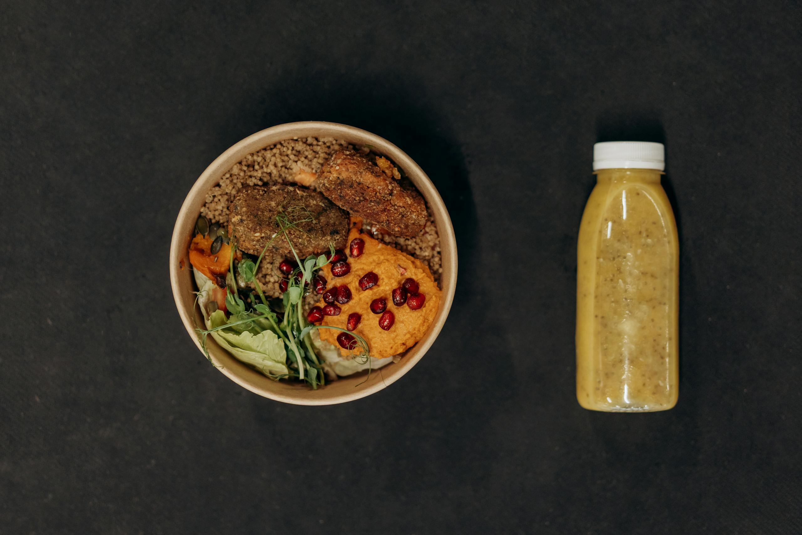 A vibrant vegan bowl with quinoa, fresh vegetables, and a smoothie bottle against a dark backdrop.