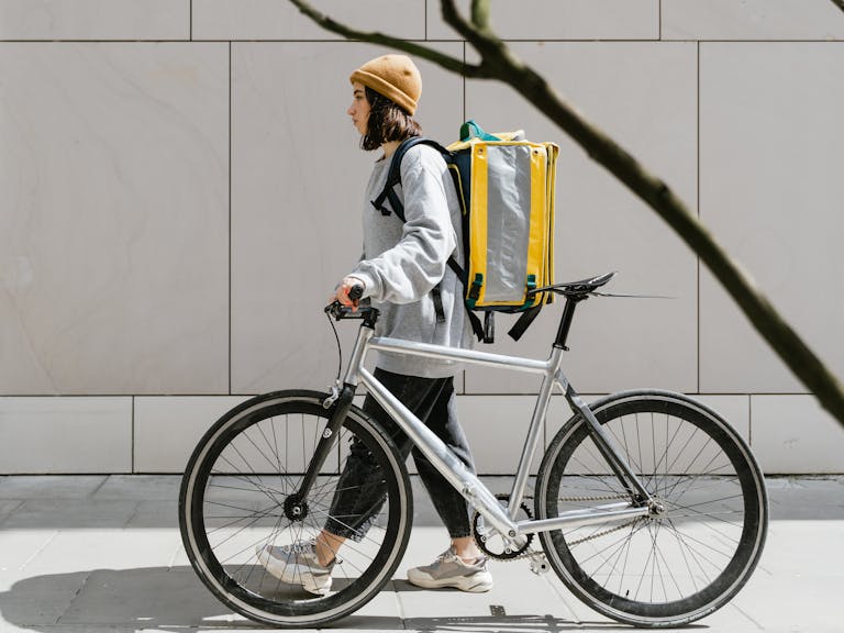 A woman walking beside a bicycle with a thermal backpack, representing urban food delivery.