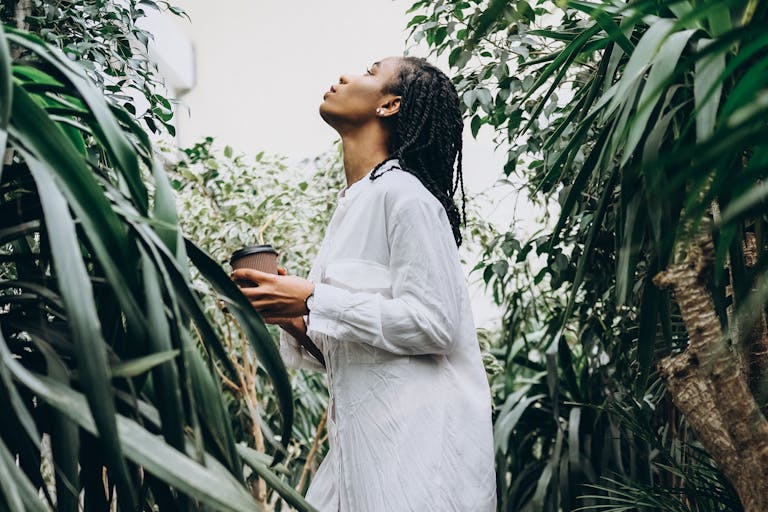 A woman with braided hair holds a coffee cup while appreciating plants in a serene greenhouse setting.