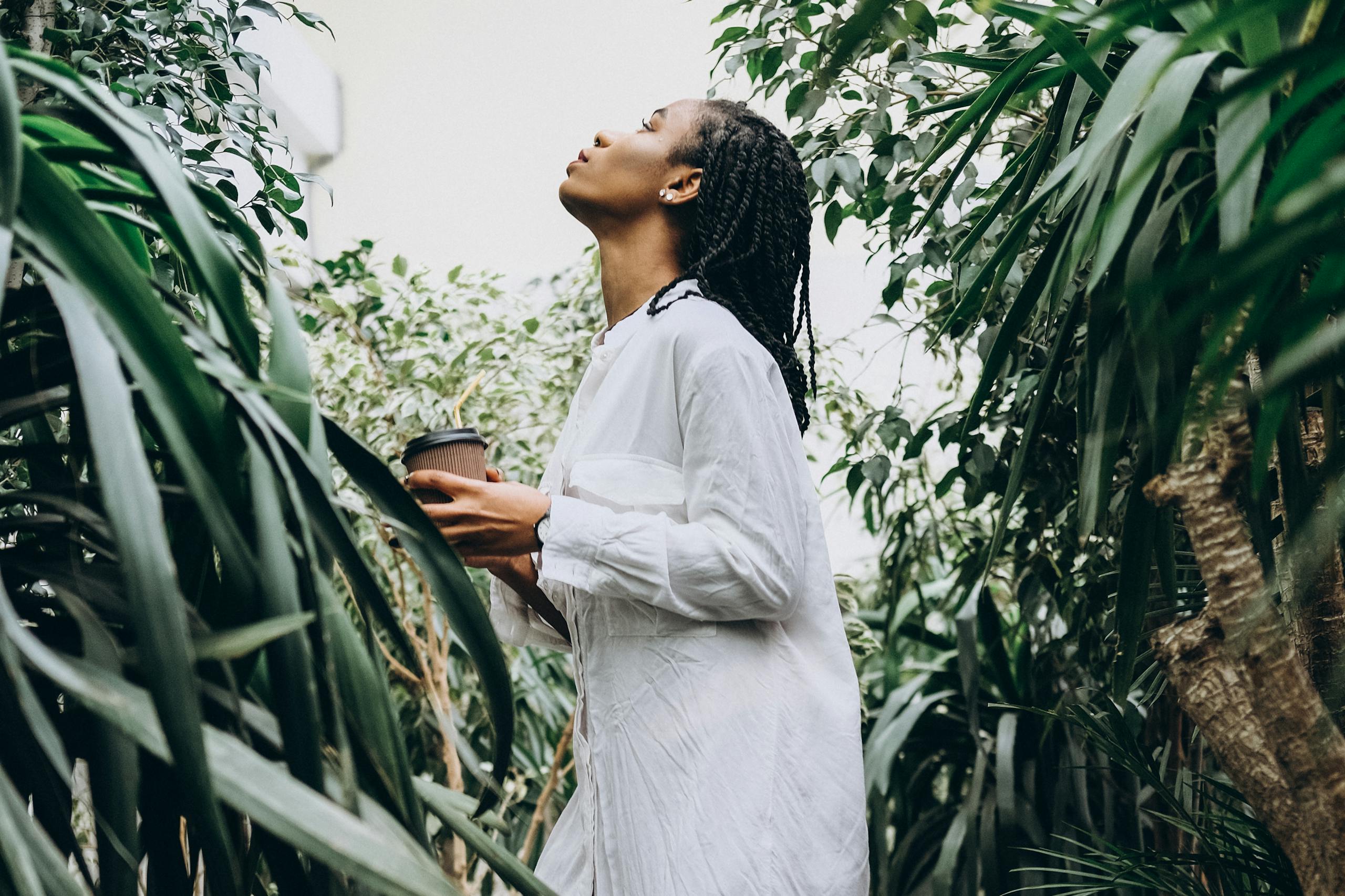A woman with braided hair holds a coffee cup while appreciating plants in a serene greenhouse setting.