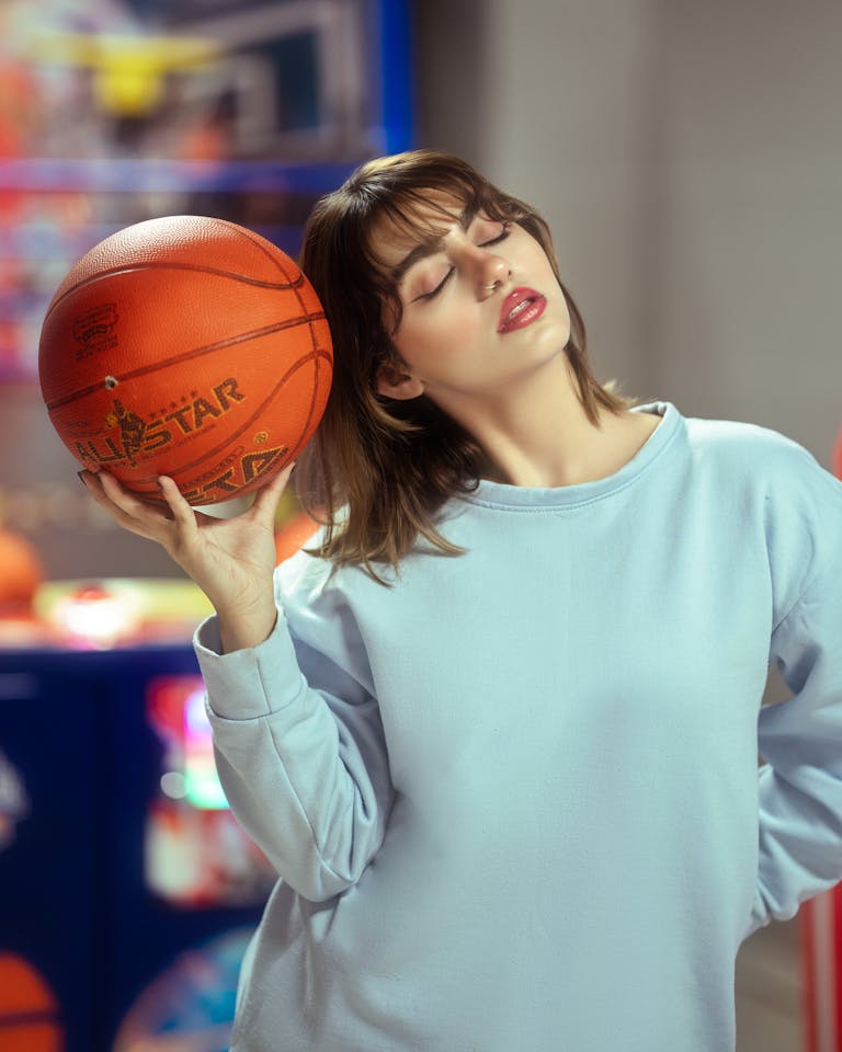 A young woman in a blue sweater holding a basketball indoors with eyes closed.