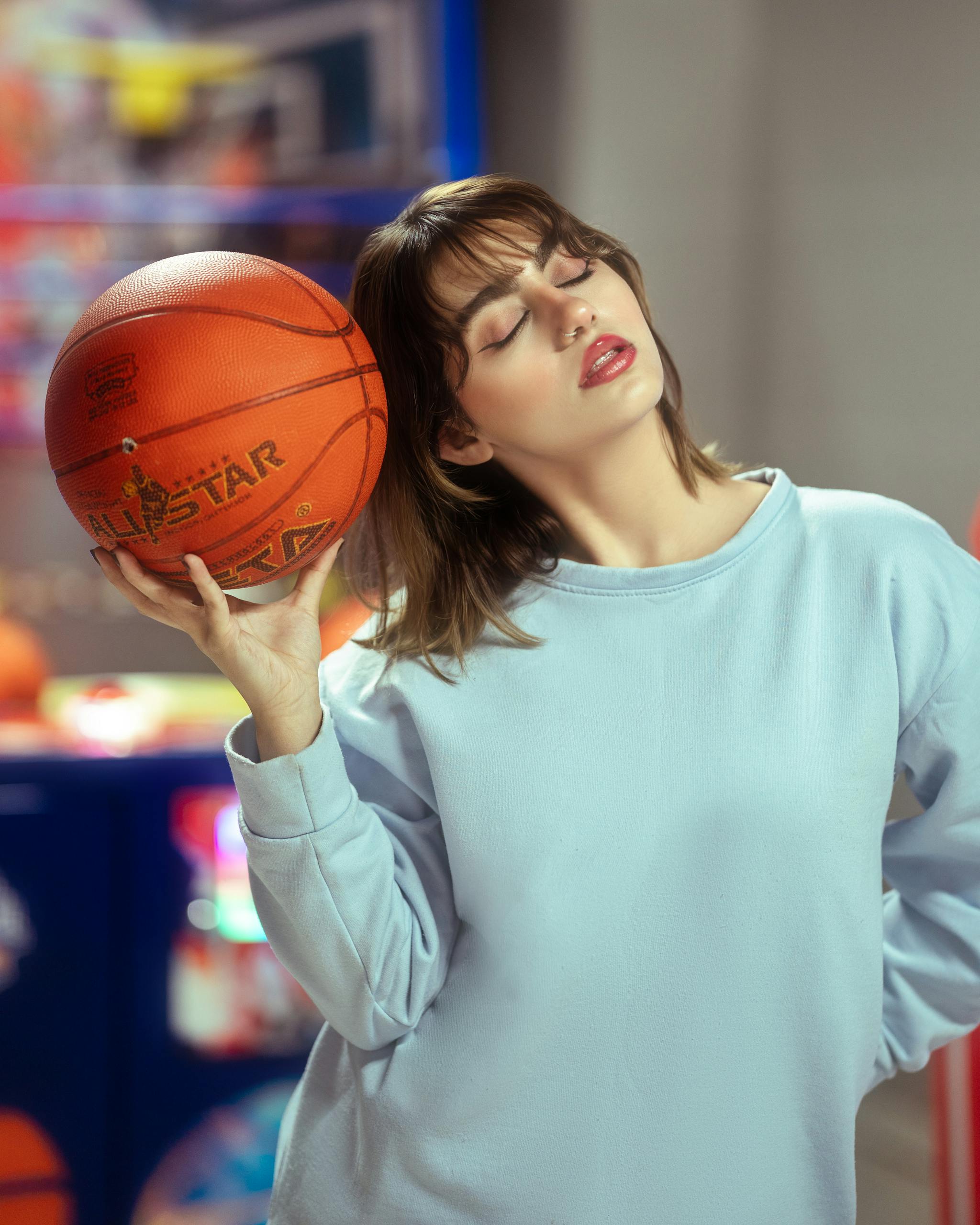 A young woman in a blue sweater holding a basketball indoors with eyes closed.