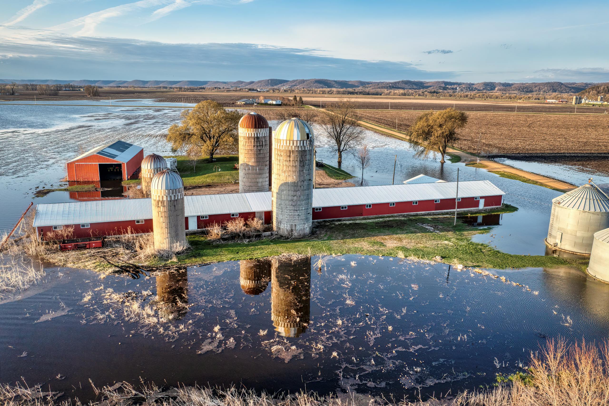 Aerial view of a flooded farm in Wabasha, MN, showcasing silos and barns in a rural landscape.