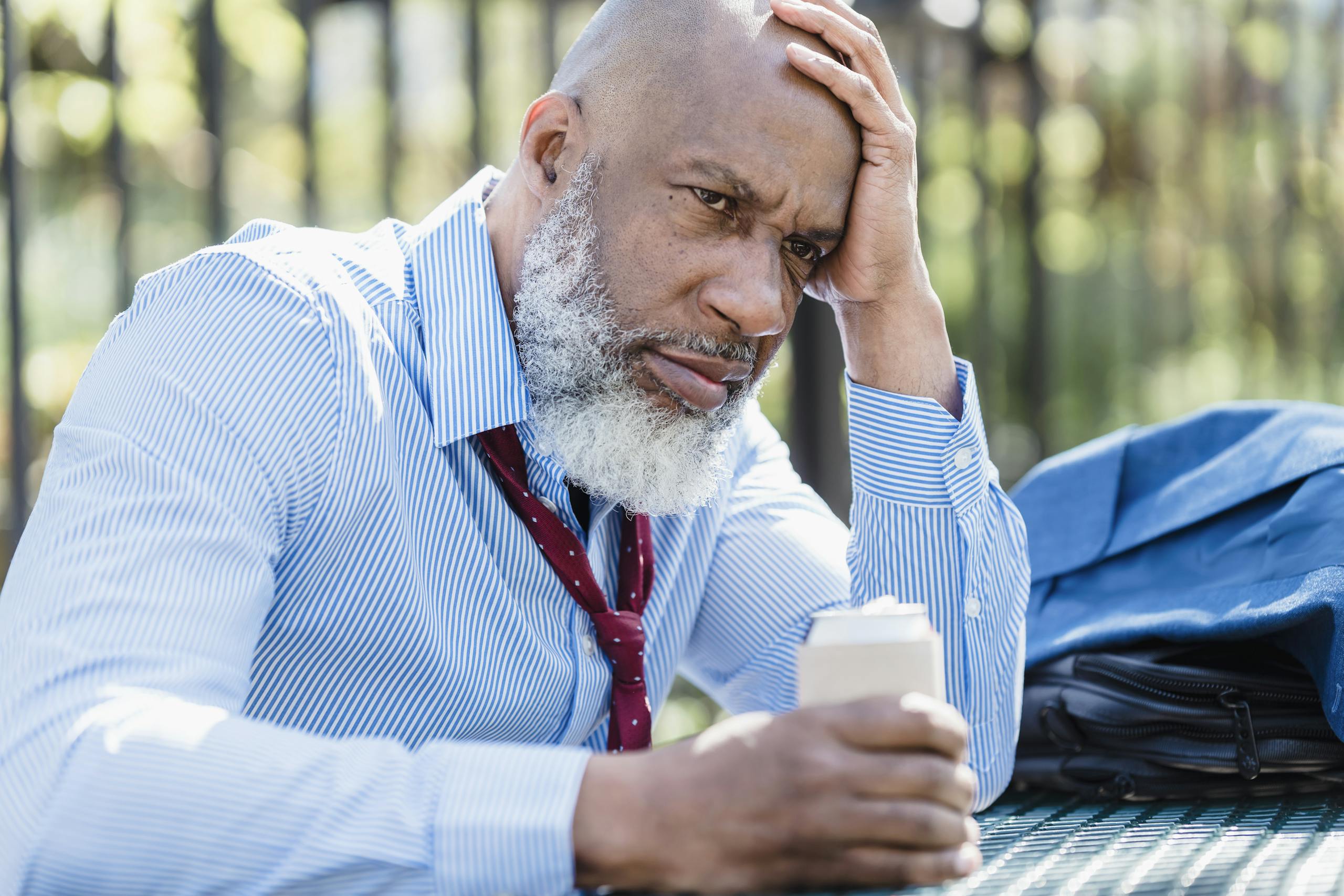 An adult man in business attire appears stressed while sitting outdoors with a drink.