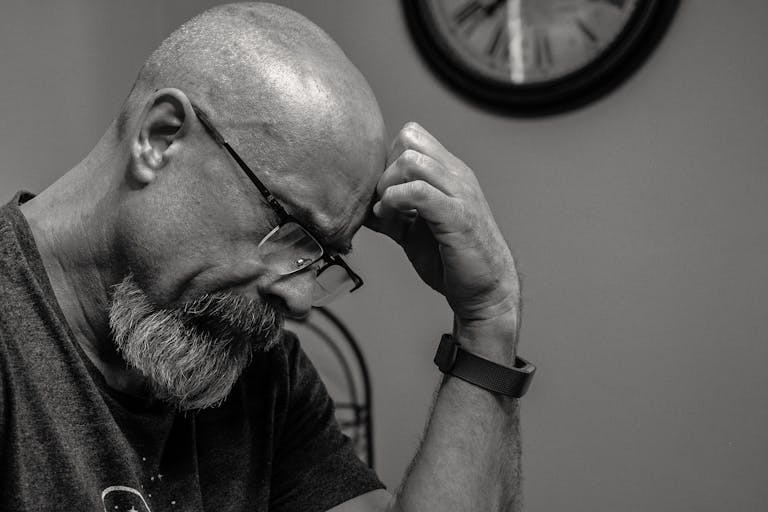 Black and white portrait of a thoughtful bald man indoors, capturing a moment of reflection with a wall clock in the background.