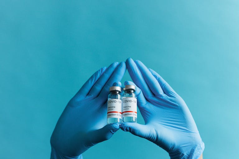 Blue gloved hands holding COVID-19 vaccine vials on a blue background.