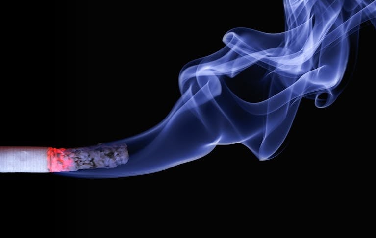 Detailed close-up of a cigarette with glowing ember and swirling smoke on a black background.