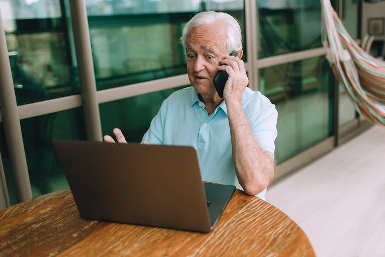 Elderly man multitasking with a laptop and phone indoors by a glass wall.