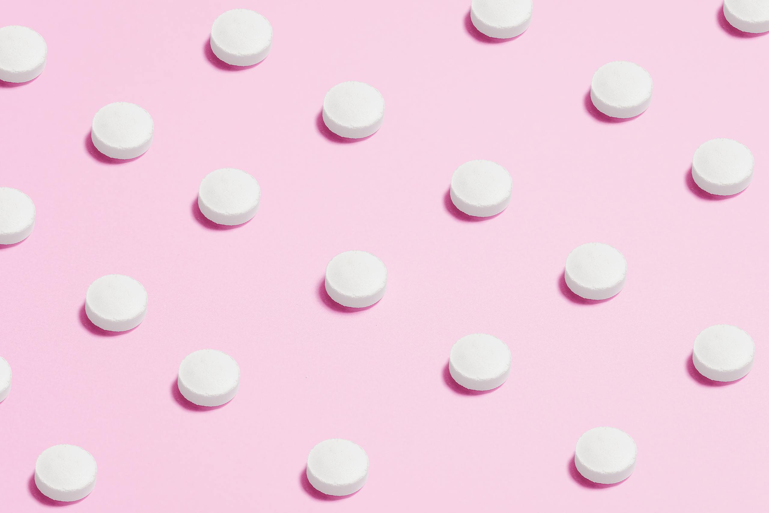 Flat lay of white pills on a pastel pink surface, showcasing a medical theme.