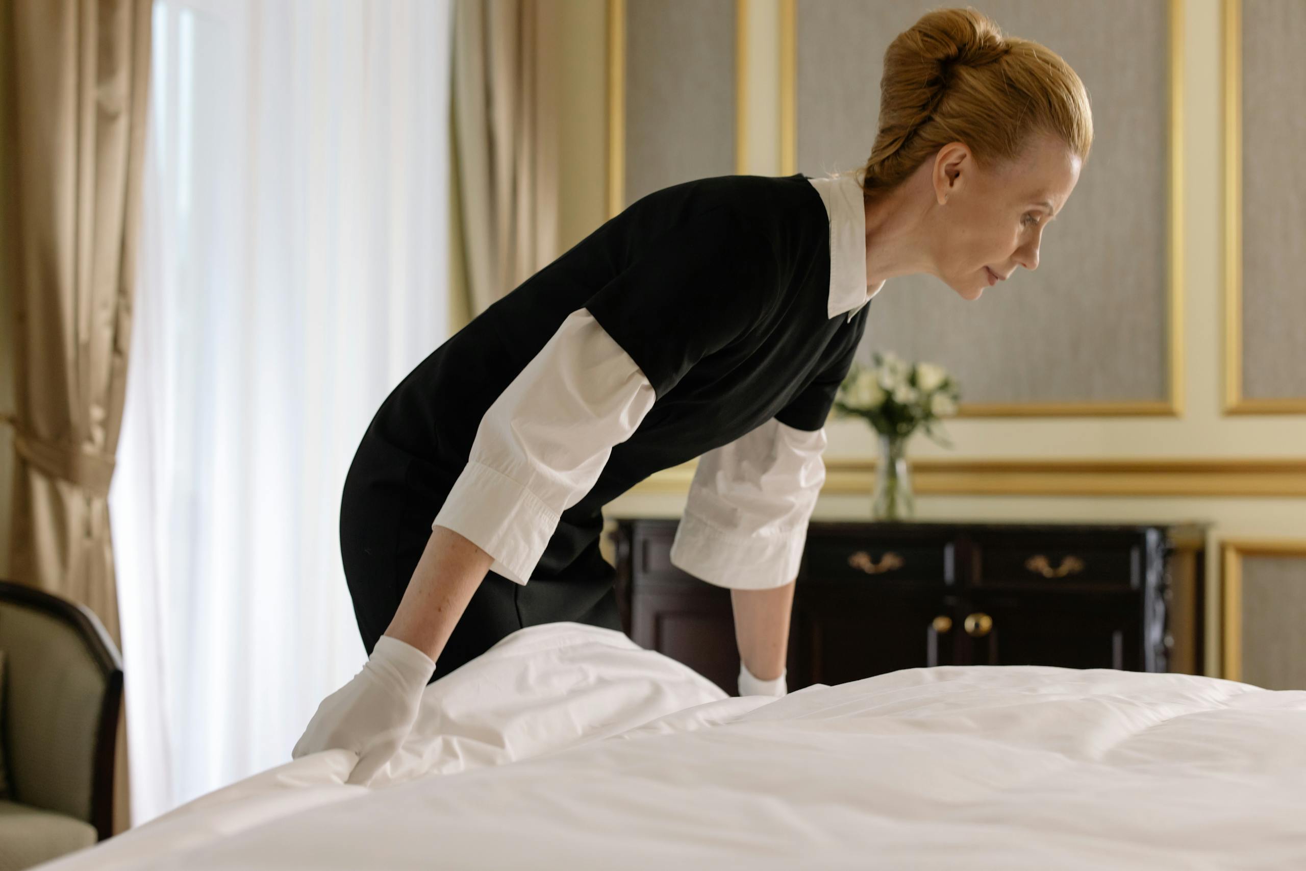 Hotel housekeeper making bed in an elegant room, wearing gloves and uniform.