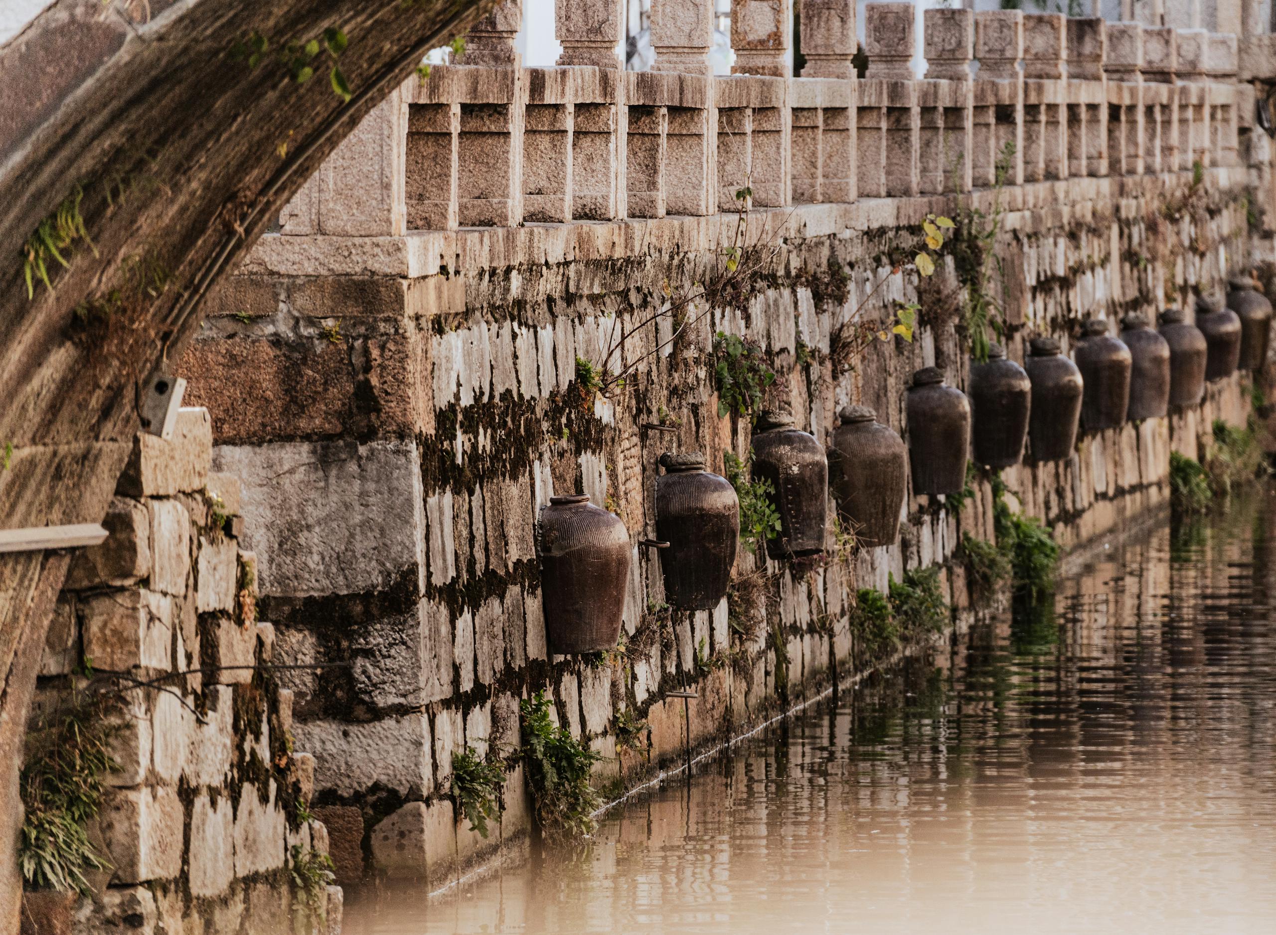 Old clay jugs line a stone wall beside a canal, showcasing rustic charm and historical ambiance.