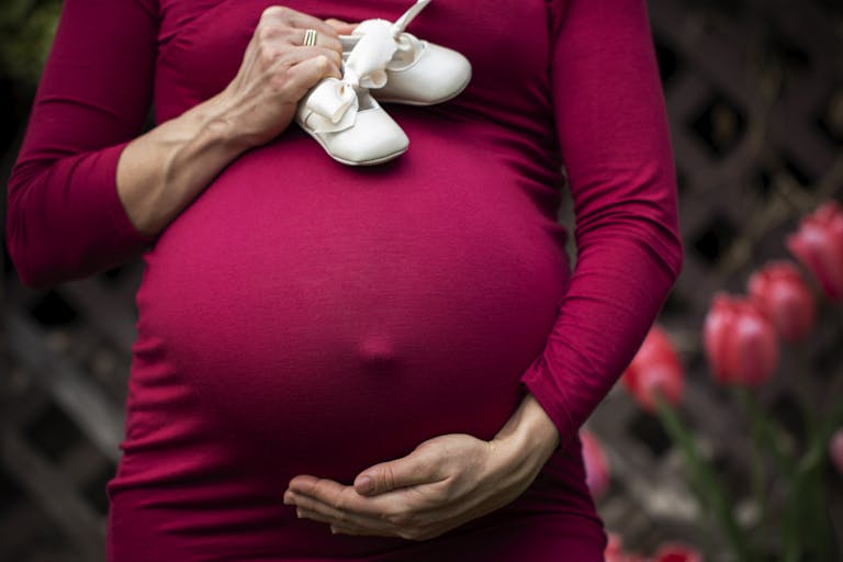 Pregnant woman holding baby shoes on belly in garden setting.