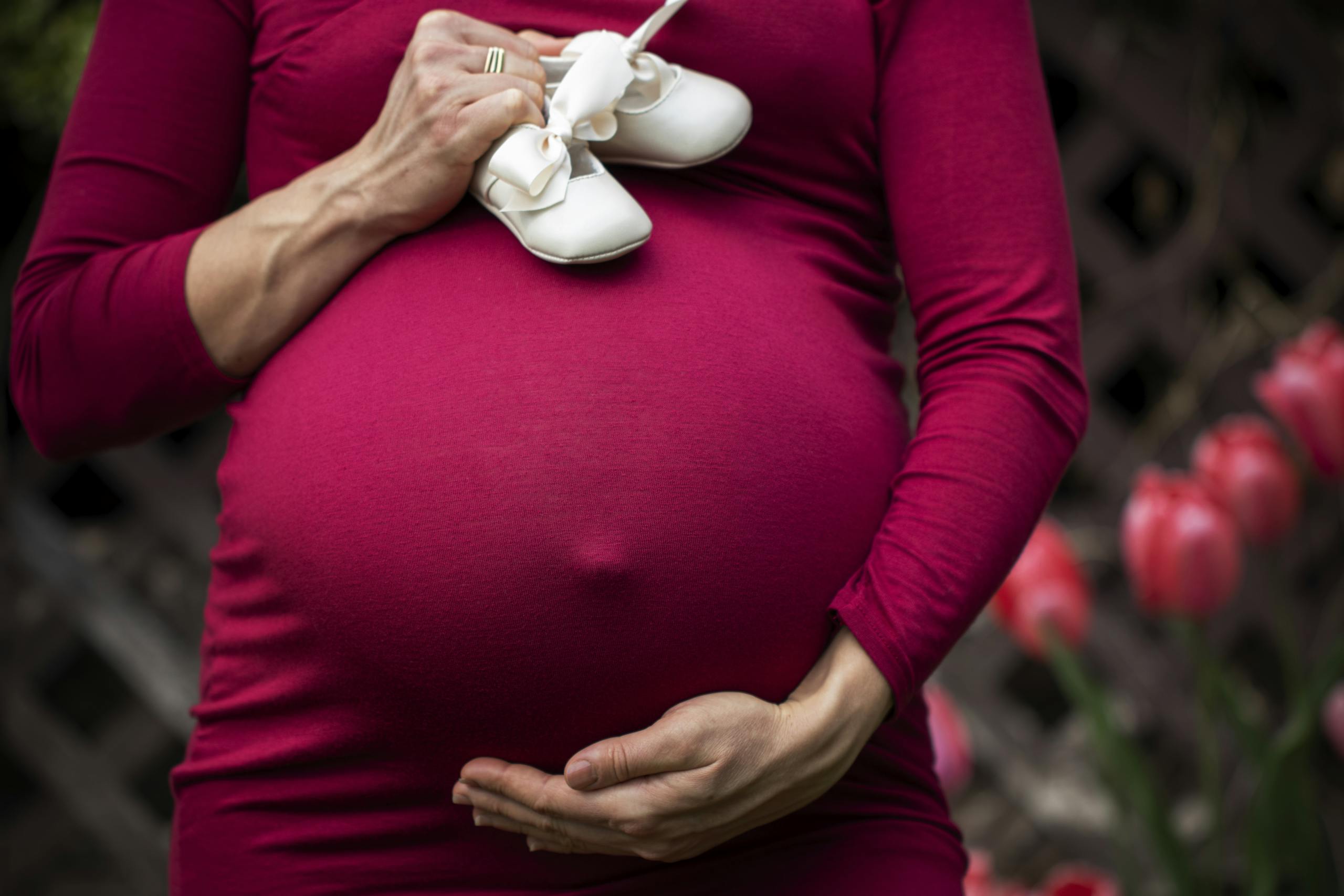 Pregnant woman holding baby shoes on belly in garden setting.