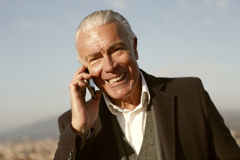 Senior man with grey hair smiling while talking on the phone outdoors with a blurred cityscape background.