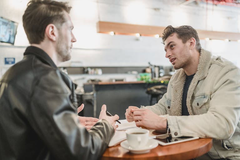 Side view of crop young man talking about business project at table with papers and looking at partner on blurred background