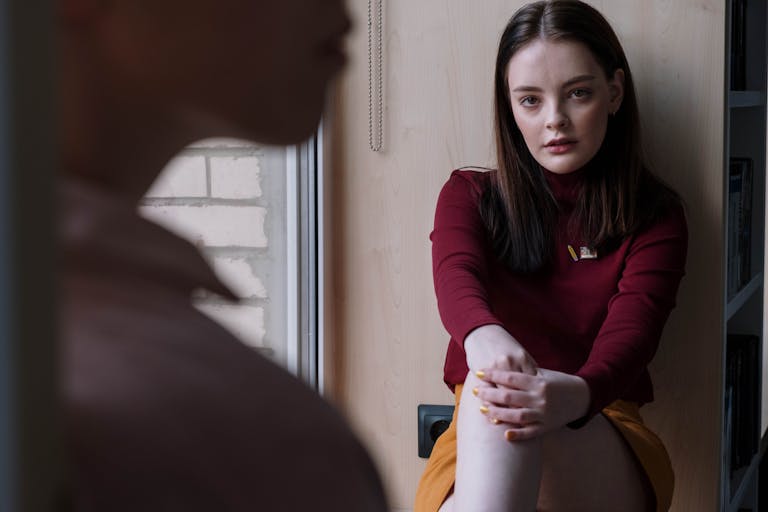 Teen girl sits by a window, lost in thought, wearing a red sweater.