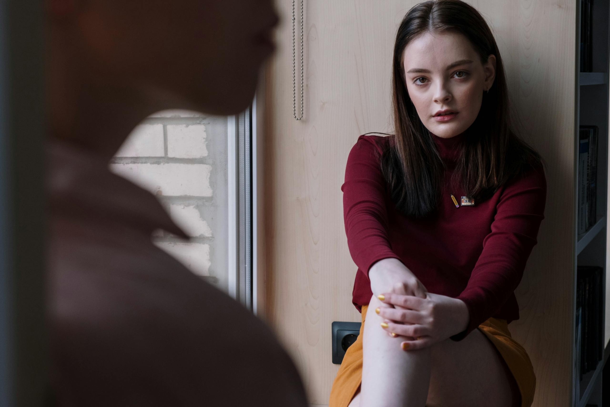 Teen girl sits by a window, lost in thought, wearing a red sweater.