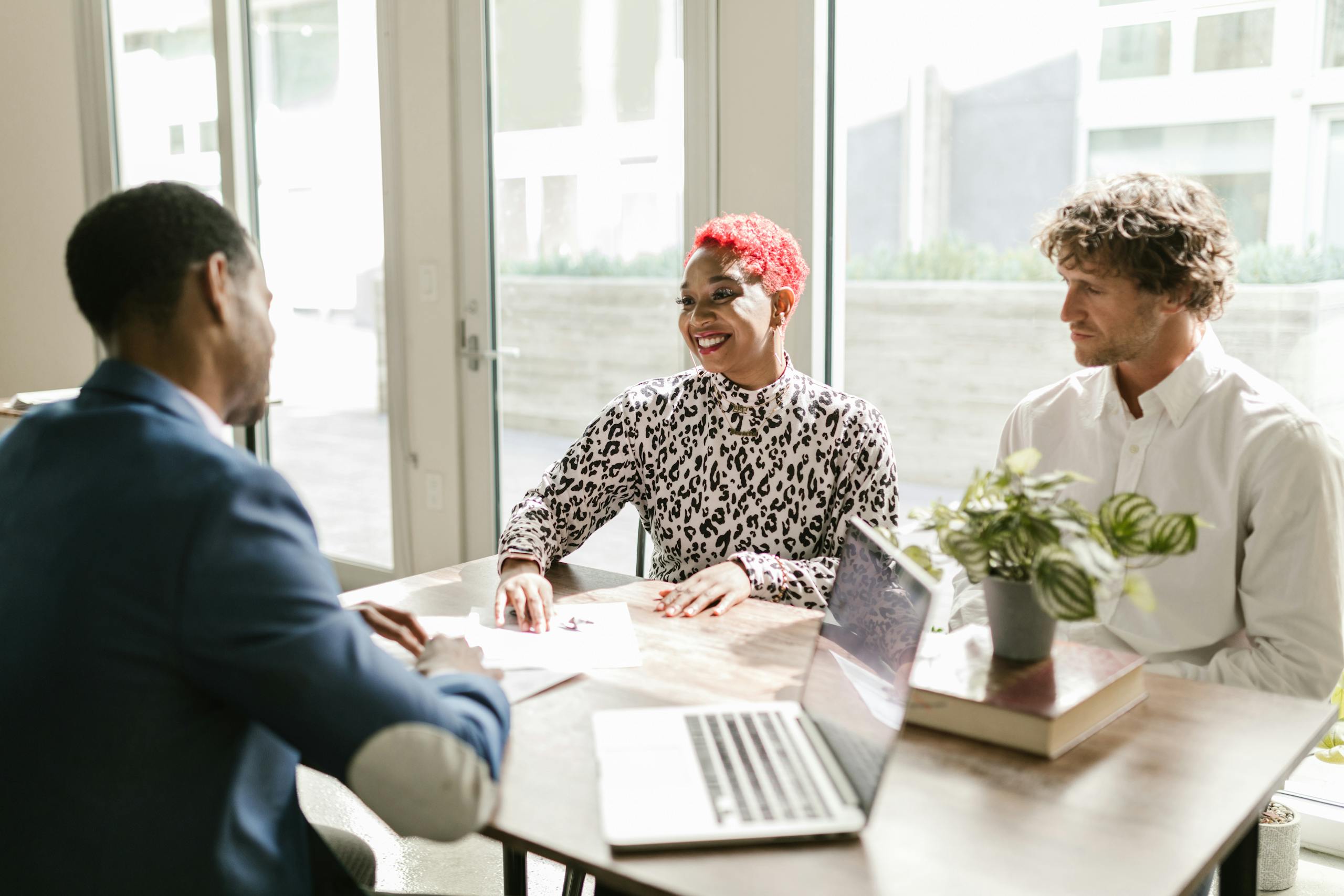Three professionals meeting in a sunny office discussing finance with smiles.