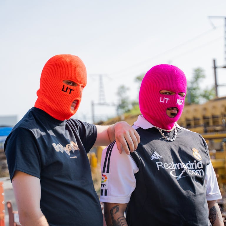 Two people in vibrant masks pose outdoors, showcasing edgy streetwear style.