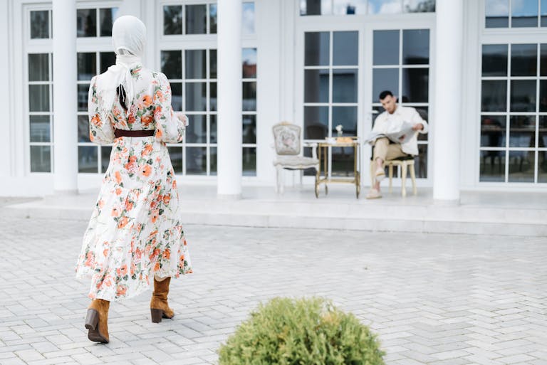 Woman in floral dress with headscarf walking towards patio with seated man.