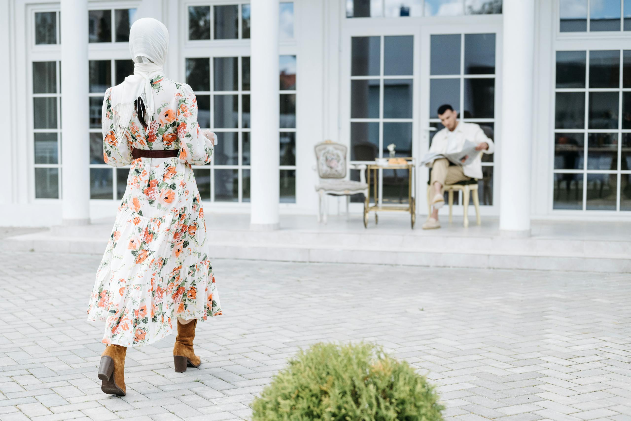 Woman in floral dress with headscarf walking towards patio with seated man.